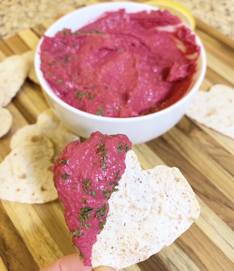 Ready to eat heart-shaped pita chip half covered with pink Athlete Beet Hummus.